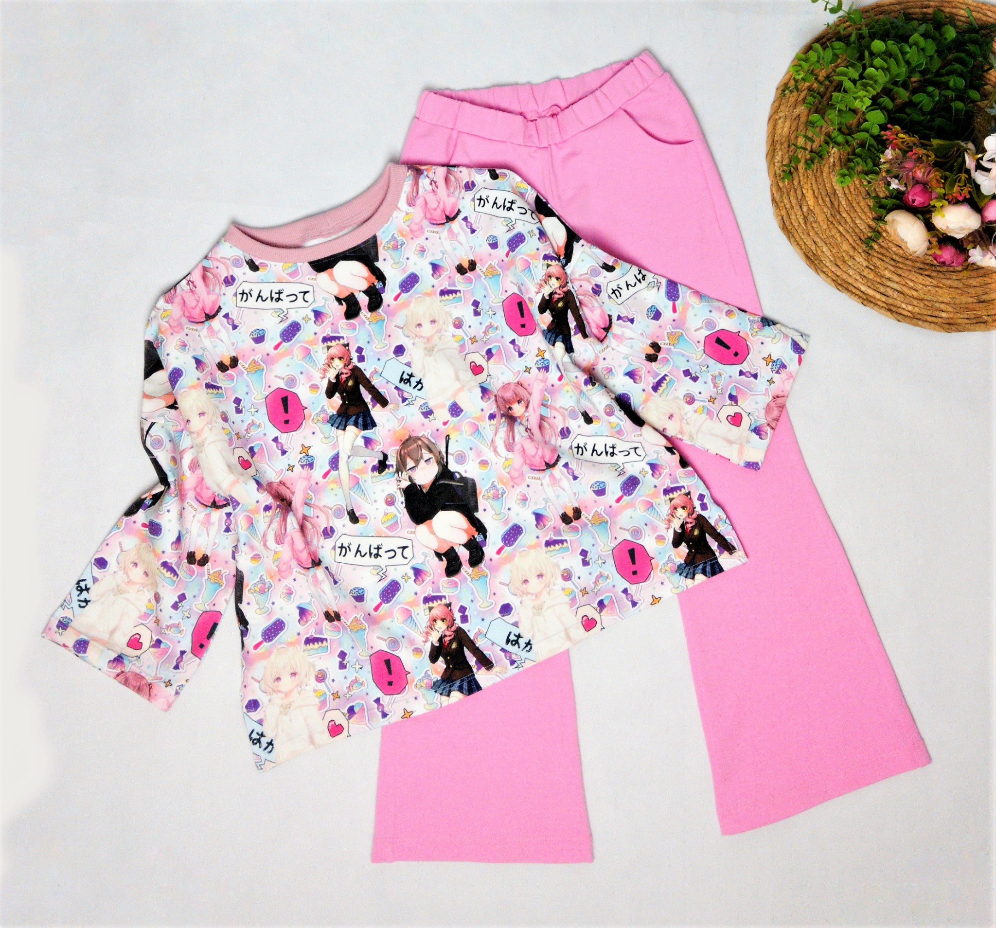 Różowa bluzka z długim rękawem i nadrukiem w stylu anime oraz dopasowane różowe spodnie, ułożone na białym tle obok dekoracyjnego koszyka z zielenią i kwiatami.