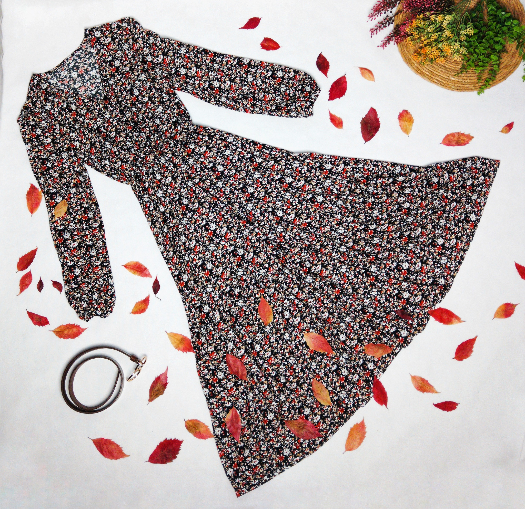 Czarna sukienka w drobne kwiaty, rozłożona na białym tle, otoczona opadłymi, czerwono-pomarańczowymi liśćmi; obok leży brązowy pasek i koszyk z kwiatami.