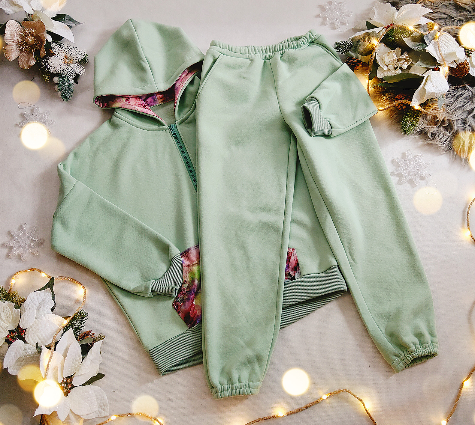 Miętowy dres dziecięcy z kapturem i kolorową podszewką, leżący na białym tle z dekoracjami świątecznymi i lampkami.