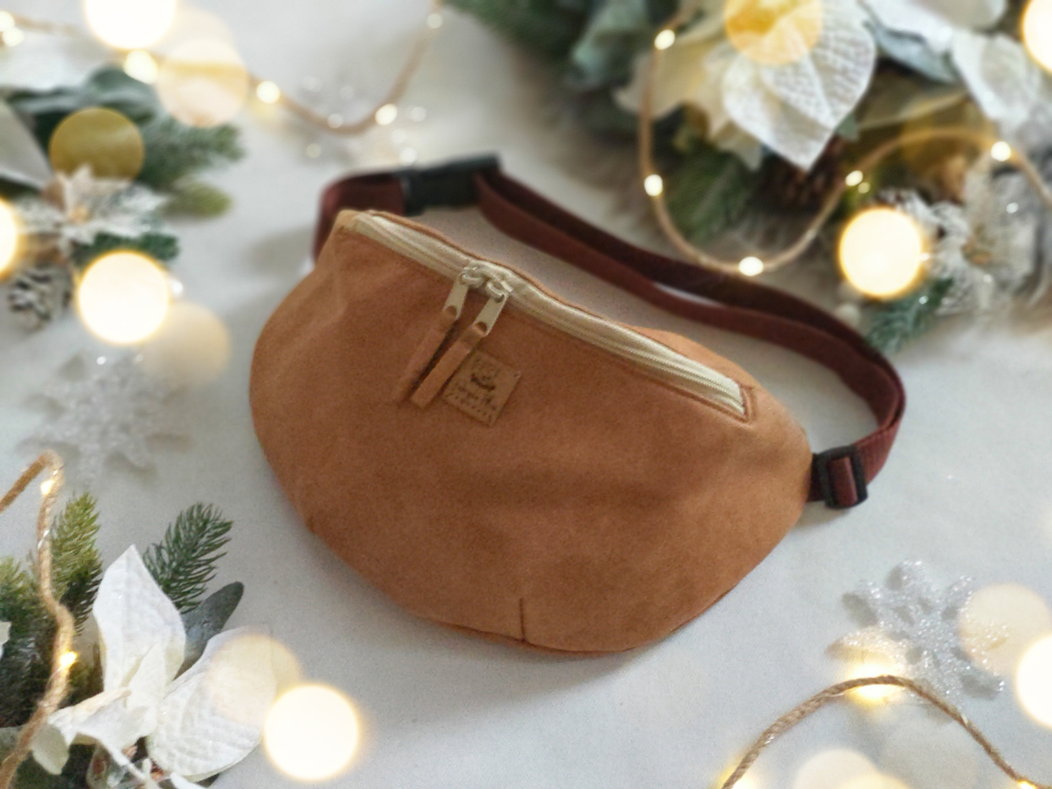 Brązowa nerka z podwójnym suwakiem i logo, leżąca na białym tle z dekoracjami świątecznymi i delikatnym oświetleniem.