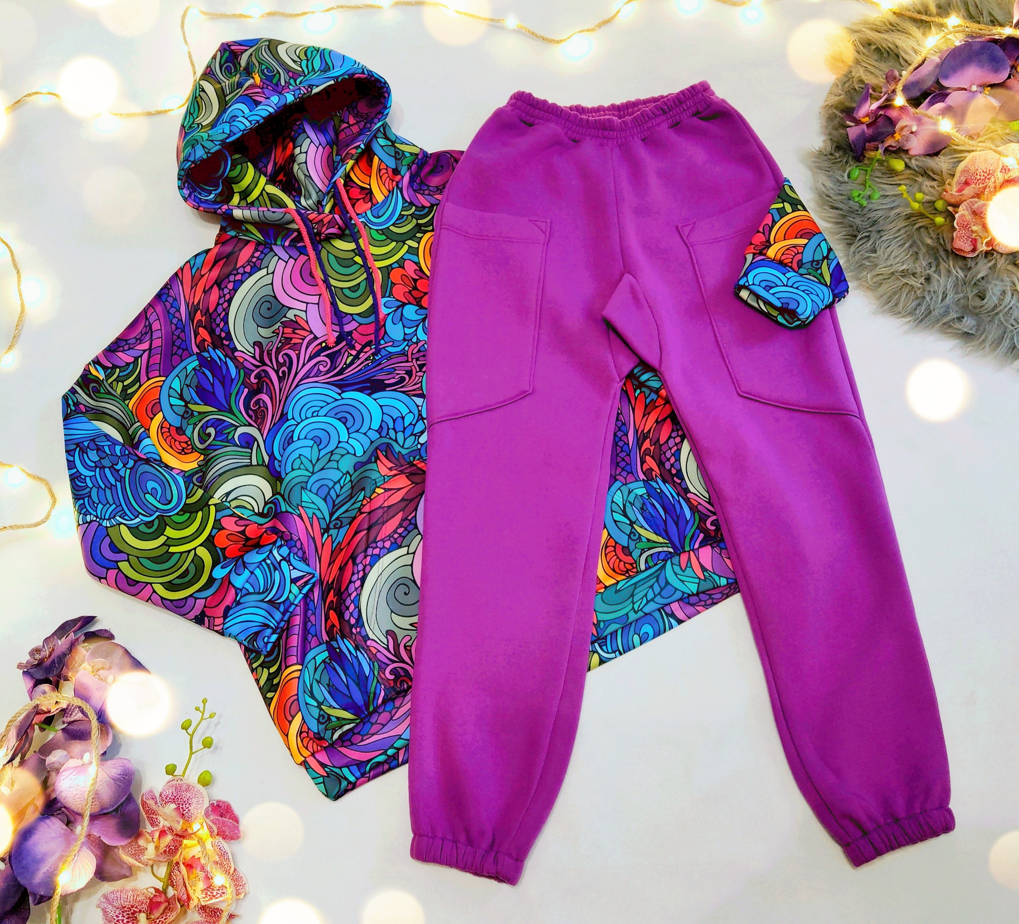 Komplet dresowy: bluza z kapturem w psychodeliczny, kolorowy wzór i fioletowe spodnie dresowe z kieszeniami, leżące na białym tle z dekoracyjnymi lampkami i kwiatami.