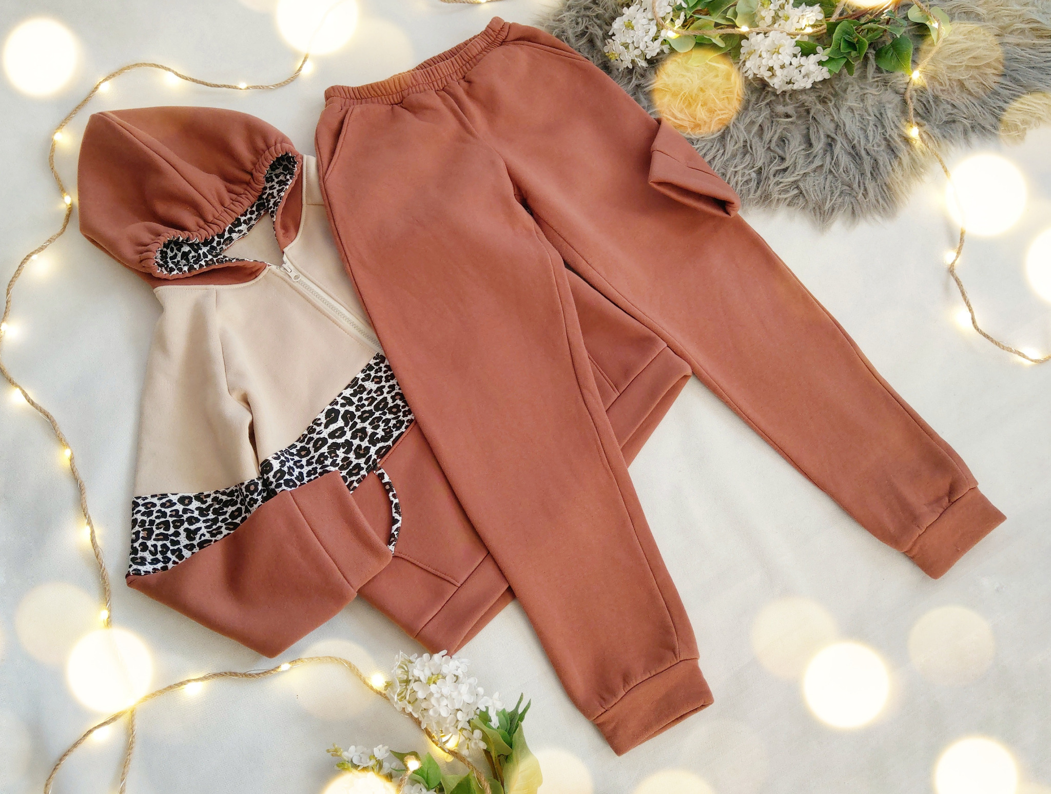 Komplet dresowy: brązowe spodnie i beżowo-brązowa bluza z kapturem, wykończona wzorem panterki, ułożone na białym tle z dekoracjami kwiatowymi i lampkami.
