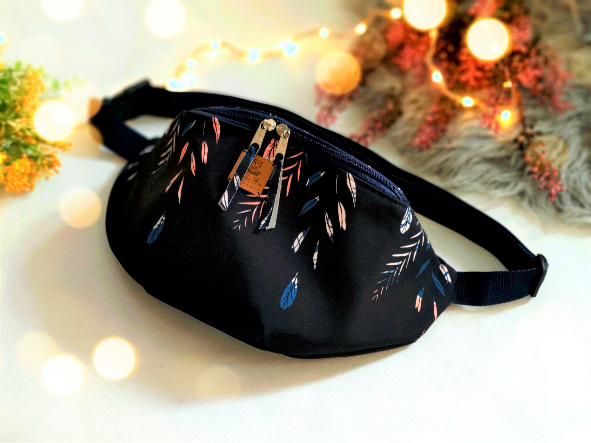 Czarna, materiałowa nerka biodrowa z motywem liści w kolorach bieli, różu i granatu, zapinana na zamek błyskawiczny z zawieszkami, leżąca na białym tle z dekoracyjnym oświetleniem i kwiatami w tle.