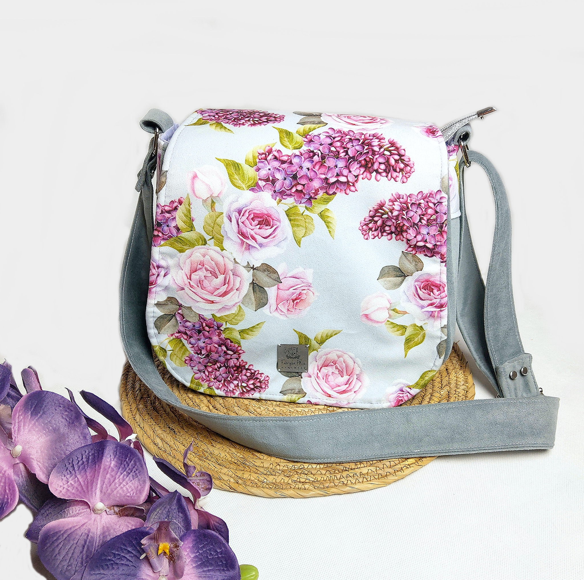Elegancka torebka damska z motywem róż i bzu, szary pasek, prezentowana na słomkowej podkładce z dekoracją z fioletowych storczyków.