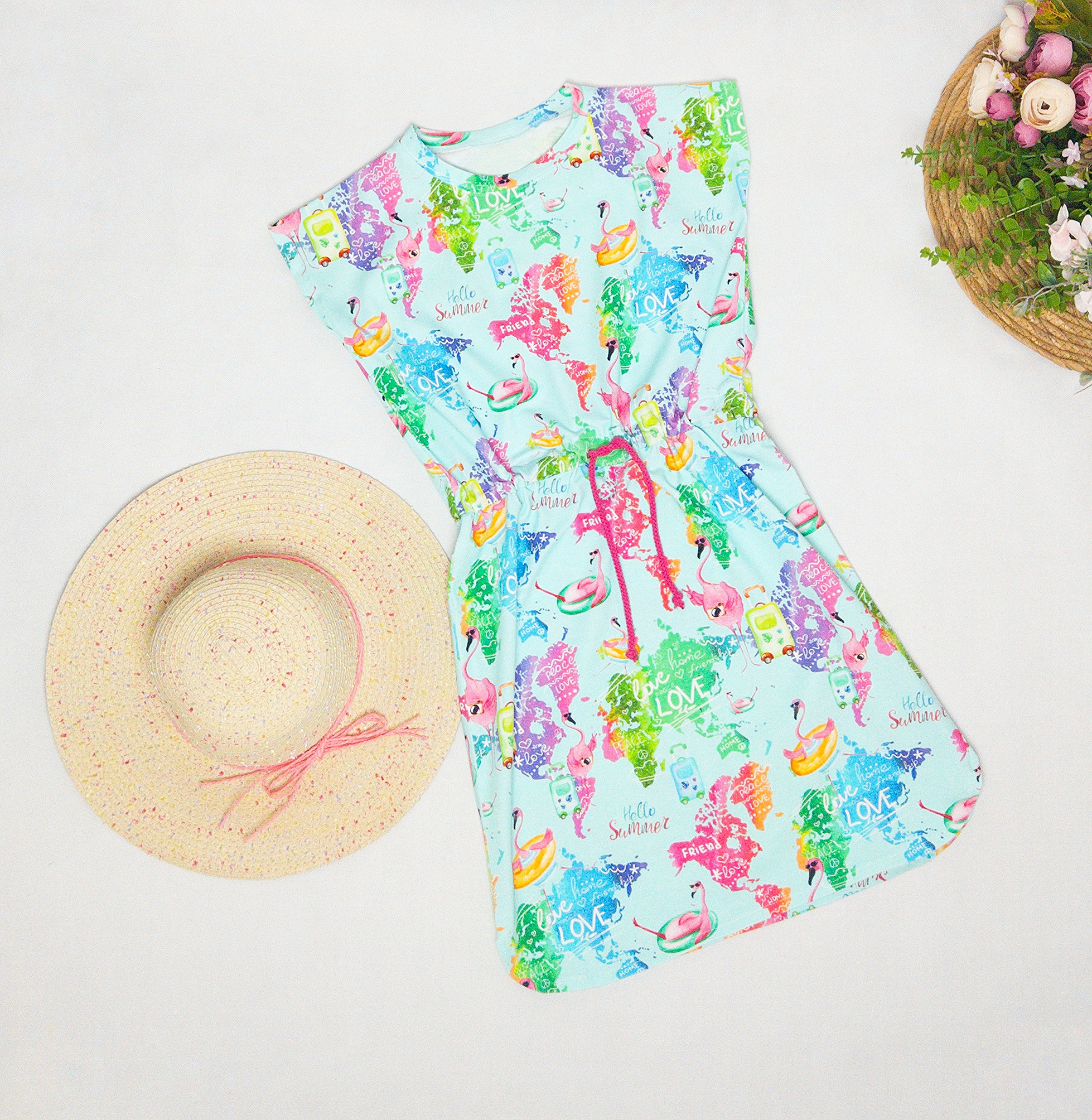 Letnia sukienka dziecięca z mapą świata, flamingami i napisami 'Hello Summer' na jasnoniebieskim tle, obok słomkowy kapelusz z różową wstążką i koszyk z kwiatami.