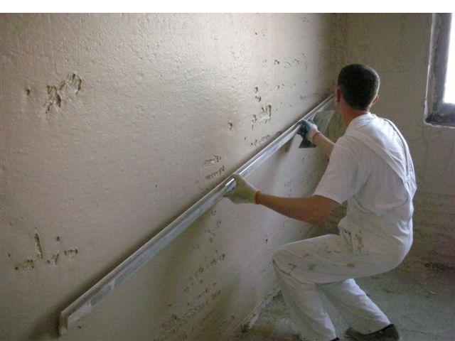 Pracownik w białym kombinezonie wygładza świeżo nałożony tynk na ścianie za pomocą długiej, aluminiowej łaty; widoczne nierówności i fragment okna w tle.