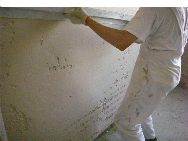 Pracownik w białym kombinezonie, z plamami tynku, wyrównuje ścianę za pomocą długiej, metalowej łaty; widoczne niedoskonałości i nierówności powierzchni przed wygładzeniem.