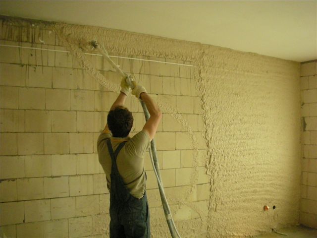 Nakładanie tynku maszynowego na ścianę z bloczków, widoczny pracownik w kombinezonie i rękawicach obsługujący lancę natryskową, fragment ściany już pokryty tynkiem.