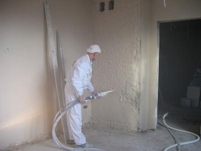 Pracownik w kombinezonie ochronnym nanosi tynk na ścianę za pomocą agregatu, widoczne węże doprowadzające materiał.