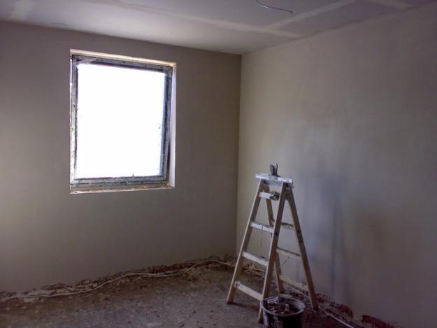 Świeżo otynkowane ściany w pomieszczeniu z oknem, widoczna drabina malarska i wiadro z narzędziami, podłoga w stanie surowym.