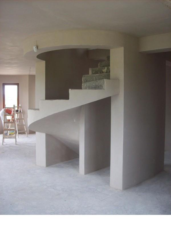 Wnętrze w trakcie prac wykończeniowych: spiralne schody z surowego betonu, ściany pokryte świeżym tynkiem, drabina oparta o ścianę w tle.