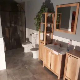 Nowoczesna łazienka z szarą glazurą, kabiną prysznicową typu walk-in, toaletą z bidetem, drewnianymi szafkami pod umywalką i wiszącą półką z roślinami.