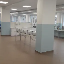 Wnętrze stołówki lub jadalni po remoncie, z jasnymi ścianami, białymi kolumnami z niebieskimi akcentami, rzędem stołów z niebieskimi krzesłami i rzędem ekspresów do kawy przy oknach.