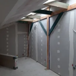 Wnętrze w trakcie montażu ścianek działowych z płyt, widoczne drewniane konstrukcje wsporcze, szpachlowane łączenia płyt, surowa podłoga i narzędzia.