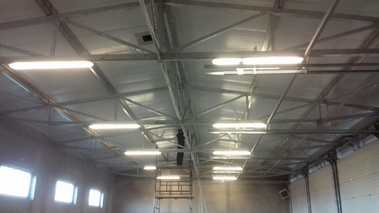 Pracownik na rusztowaniu montuje oświetlenie jarzeniowe pod metalową konstrukcją dachu hali przemysłowej.