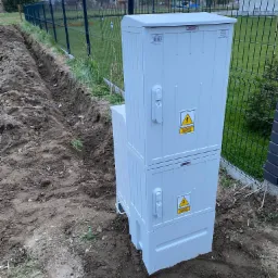 Dwu-segmentowa, biała szafa elektryczna z żółtymi znakami ostrzegawczymi 'Nie dotykać! Grozi porażeniem elektrycznym' umieszczona na ziemi przy wykopanym rowie i metalowym ogrodzeniu w tle.