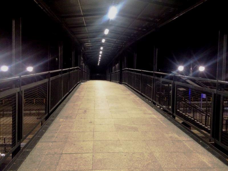 Zadaszone przejście dla pieszych nad torami kolejowymi, oświetlone rzędem lamp, z metalową konstrukcją i wyłożone płytkami chodnikowymi.