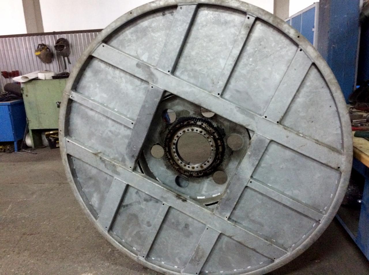 Metalowa szpula konstrukcyjna z centralnym otworem i wzmocnieniami, widoczne ślady użytkowania i zabrudzenia, w tle warsztat.