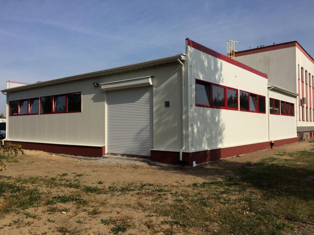 Nowoczesna hala magazynowa z szarą bramą rolowaną i czerwonymi ramami okiennymi, widok z zewnątrz na tle budynku.