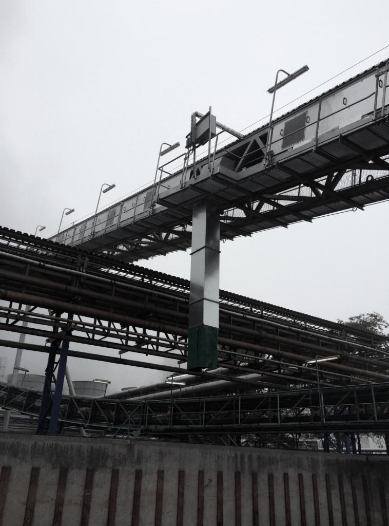 Zewnętrzny stalowy pomost technologiczny z oświetleniem, wsparty na metalowej kolumnie, nad rurociągami przemysłowymi, w pochmurny dzień.