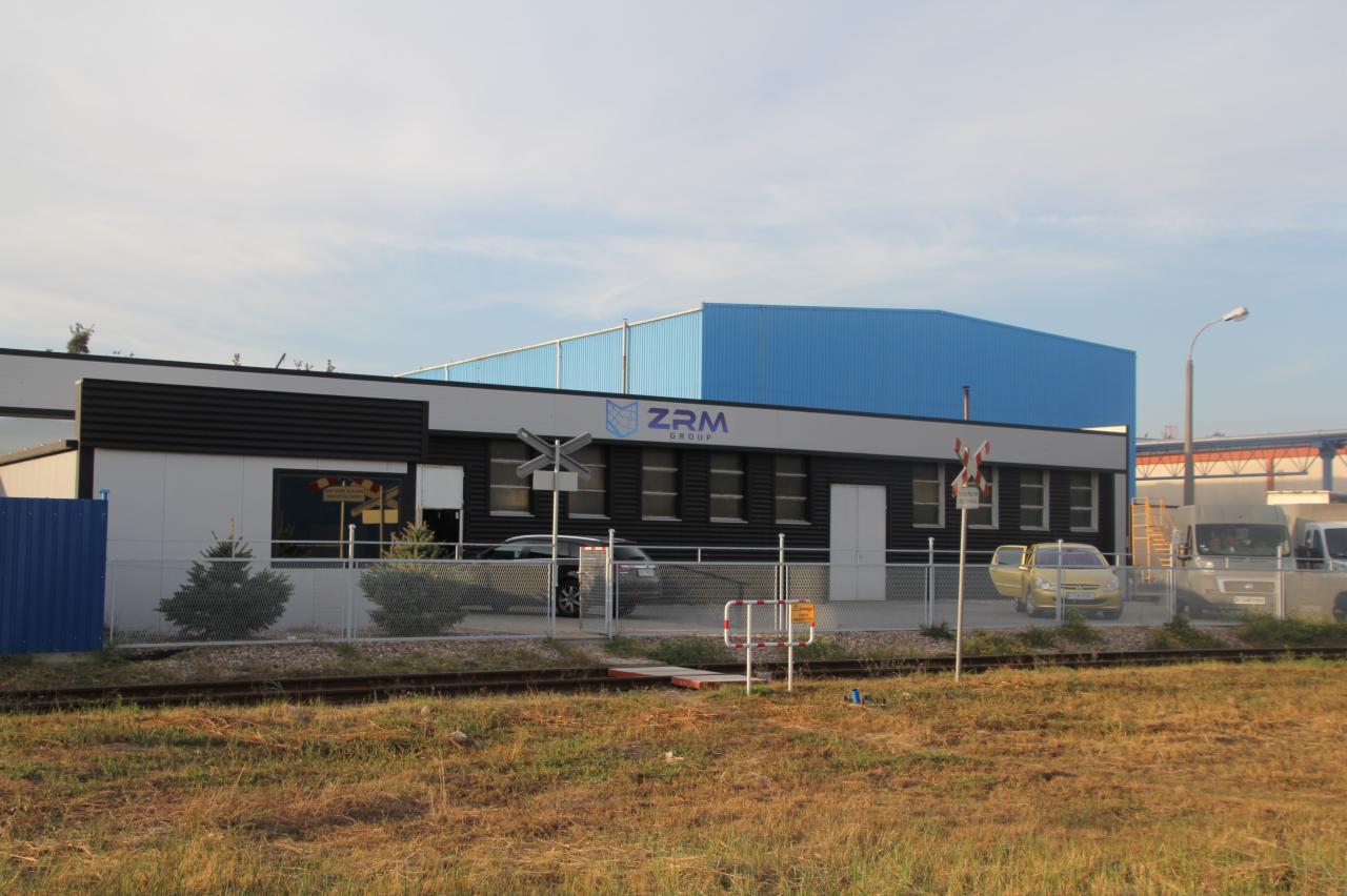 Budynek biurowy z logo ZRM Group przylegający do hali produkcyjnej z niebieskim dachem, widok z poziomu torów kolejowych.