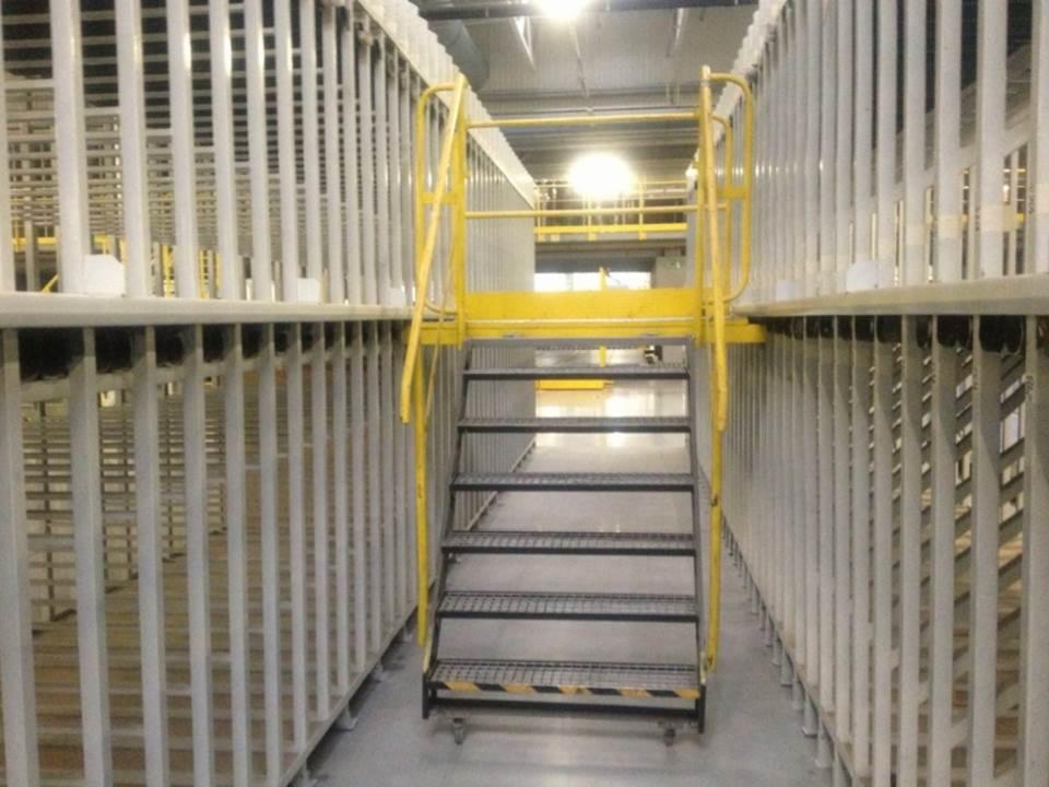 Wnętrze hali magazynowej z regałami paletowymi i schodami dostępowymi w kolorze żółtym.