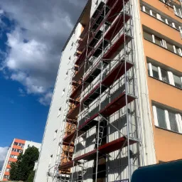 Wysoki blok z częściowo zamontowanym rusztowaniem, widok z dołu, ukazujący białą i pomarańczową elewację oraz fragment nieba z chmurami.