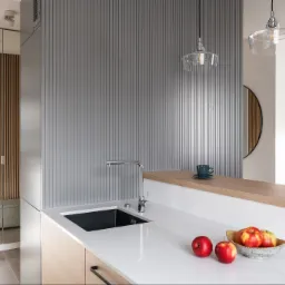 Nowoczesna kuchnia z białym blatem, czarnym zlewem i chromowaną baterią, uzupełniona o drewniane akcenty, szarą ścianę z pionowymi żłobieniami oraz dekoracyjną misę z jabłkami na blacie.