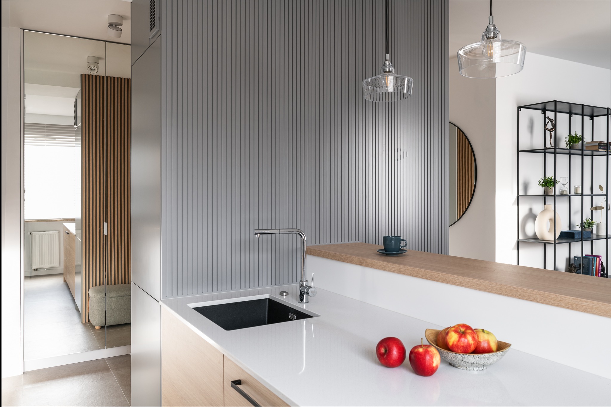 Nowoczesna kuchnia z białym blatem, czarnym zlewem i chromowaną baterią, uzupełniona o drewniane akcenty, szarą ścianę z pionowymi żłobieniami oraz dekoracyjną misę z jabłkami na blacie.