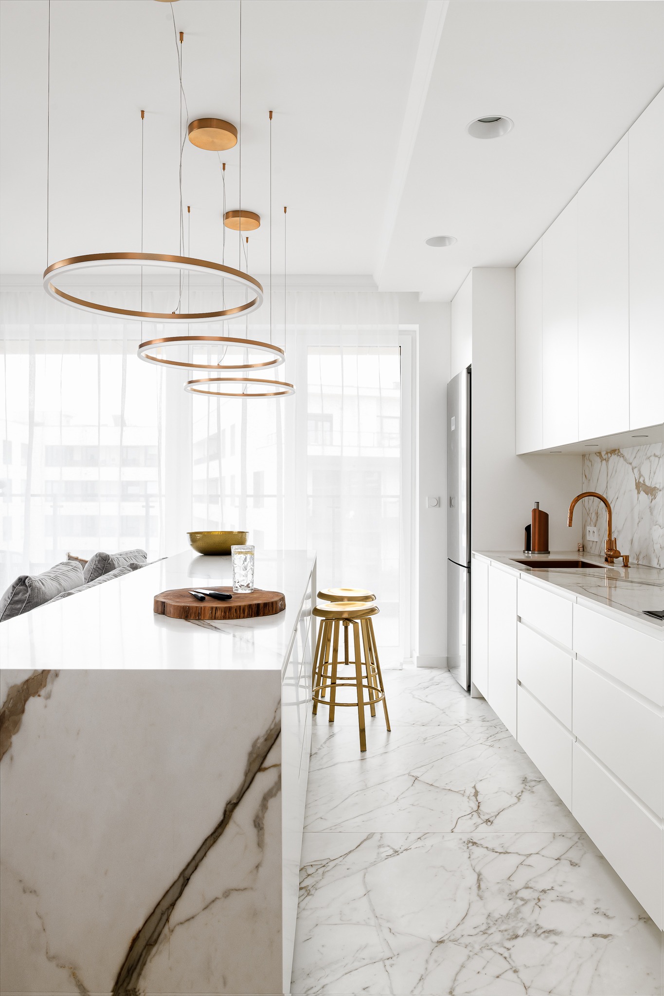 Nowoczesna kuchnia: białe szafki, marmurowy blat i podłoga z żyłkowaniem, złote hokery i okrągłe lampy wiszące.
