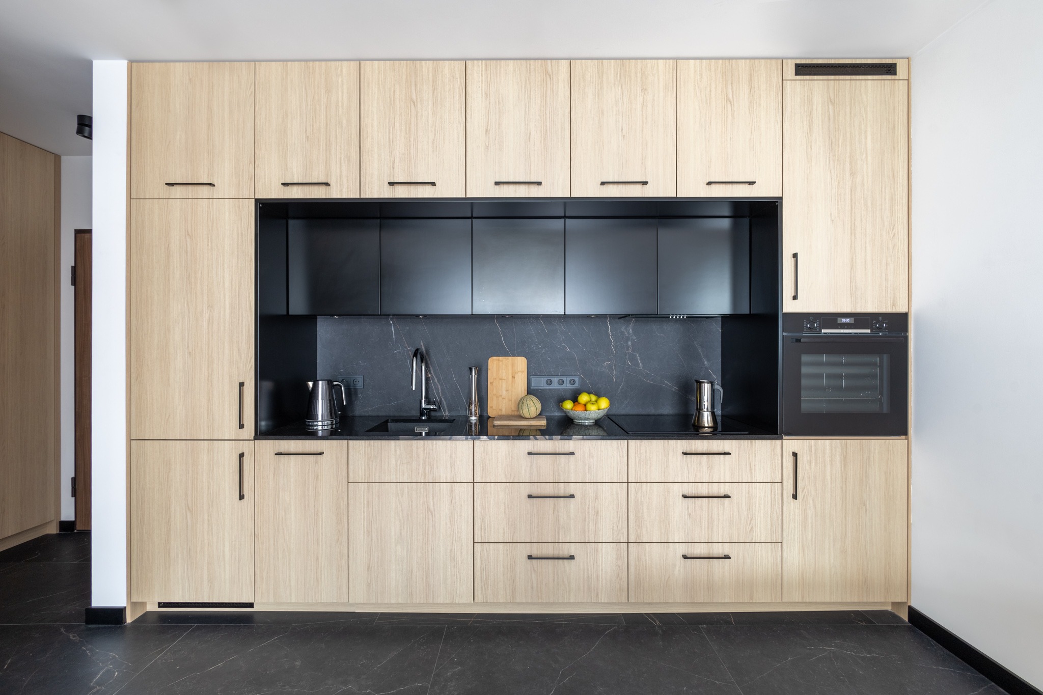 Nowoczesna kuchnia z frontami w jasnym drewnie i czarnym blatem z marmurowym wzorem, minimalistyczne uchwyty, owoce w misce na blacie.