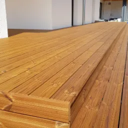 Nowy, drewniany taras z widocznymi schodkami i jasną elewacją budynku w tle. Widoczne ślady montażu na brzegach tarasu.