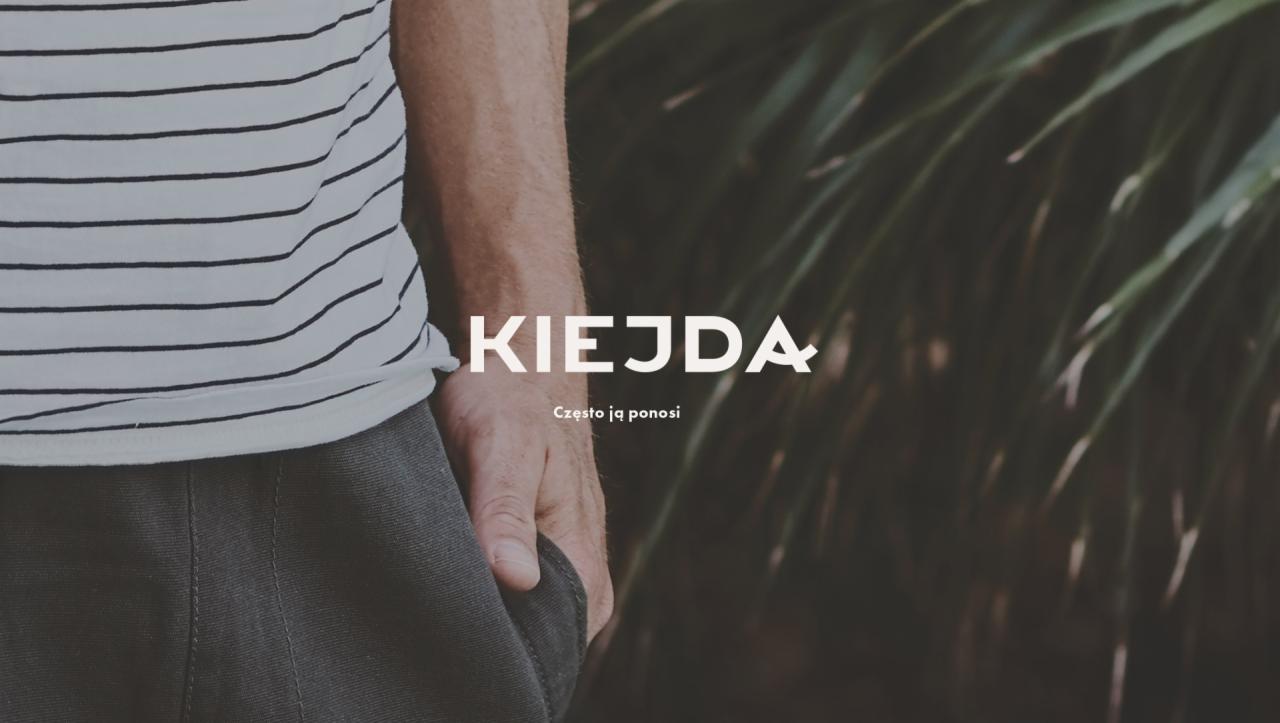 Zdjęcie przedstawia fragment osoby w paski koszulce i szarych spodniach, z dłonią w kieszeni na tle zielonych liści, z nałożonym tekstem 'KIEJDA Często ją ponosi'.