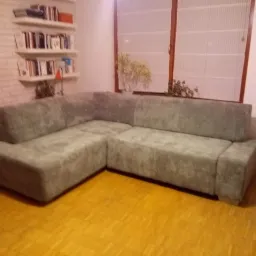 Szara narożna sofa z nową tapicerką w salonie z drewnianą podłogą, na tle ceglanej ściany z półkami na książki i okna.