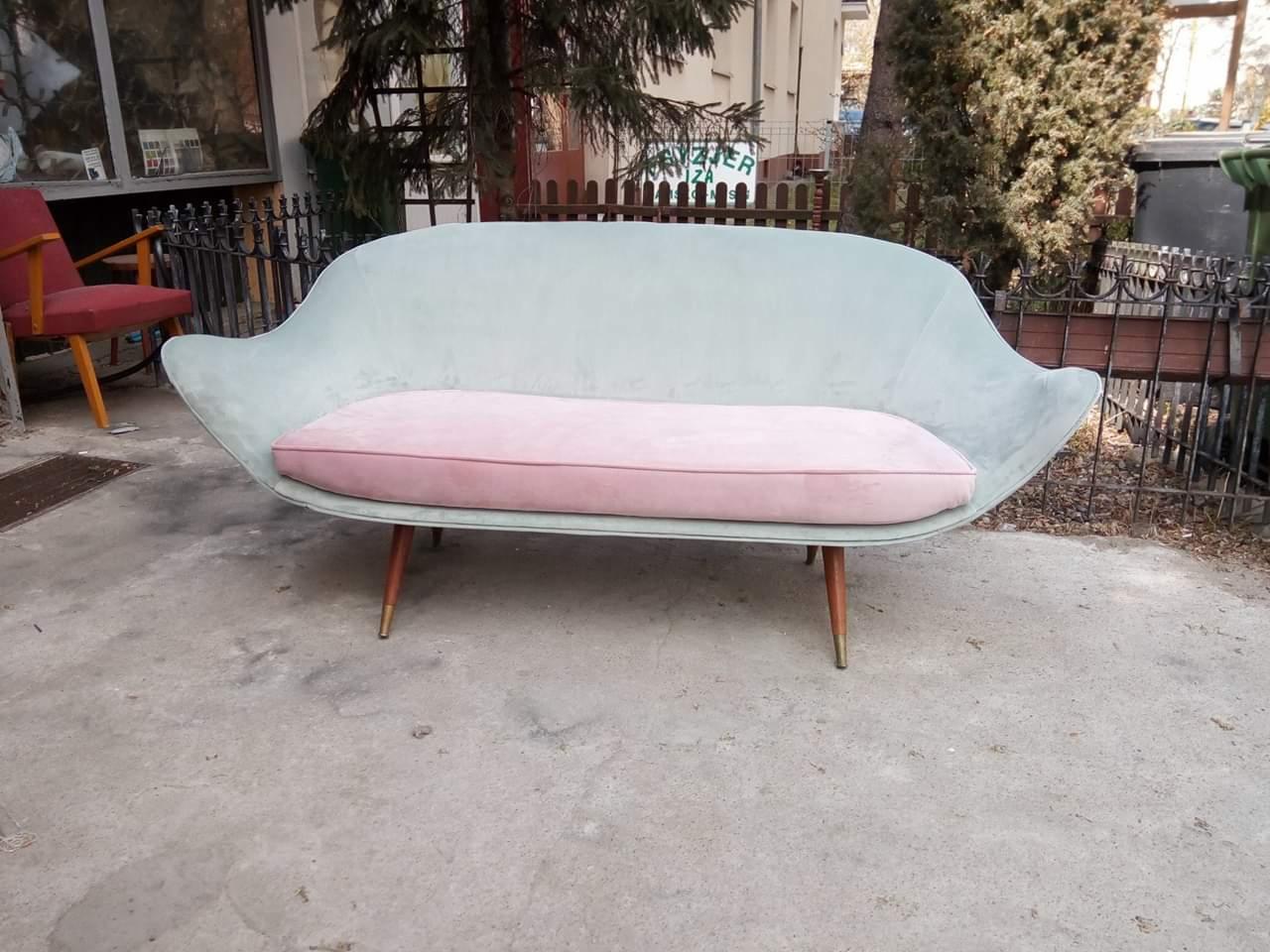 Odrestaurowana sofa w stylu vintage z jasnoniebieską tapicerką i różową poduszką siedziska, ustawiona na zewnątrz na betonowym podłożu, z widocznym fragmentem metalowego ogrodzenia w tle.