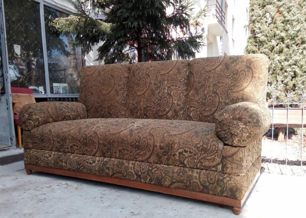 Trzyosobowa sofa z brązowym wzorem paisley, ustawiona na zewnątrz na betonowej powierzchni, z widocznymi drzewami i budynkiem w tle.