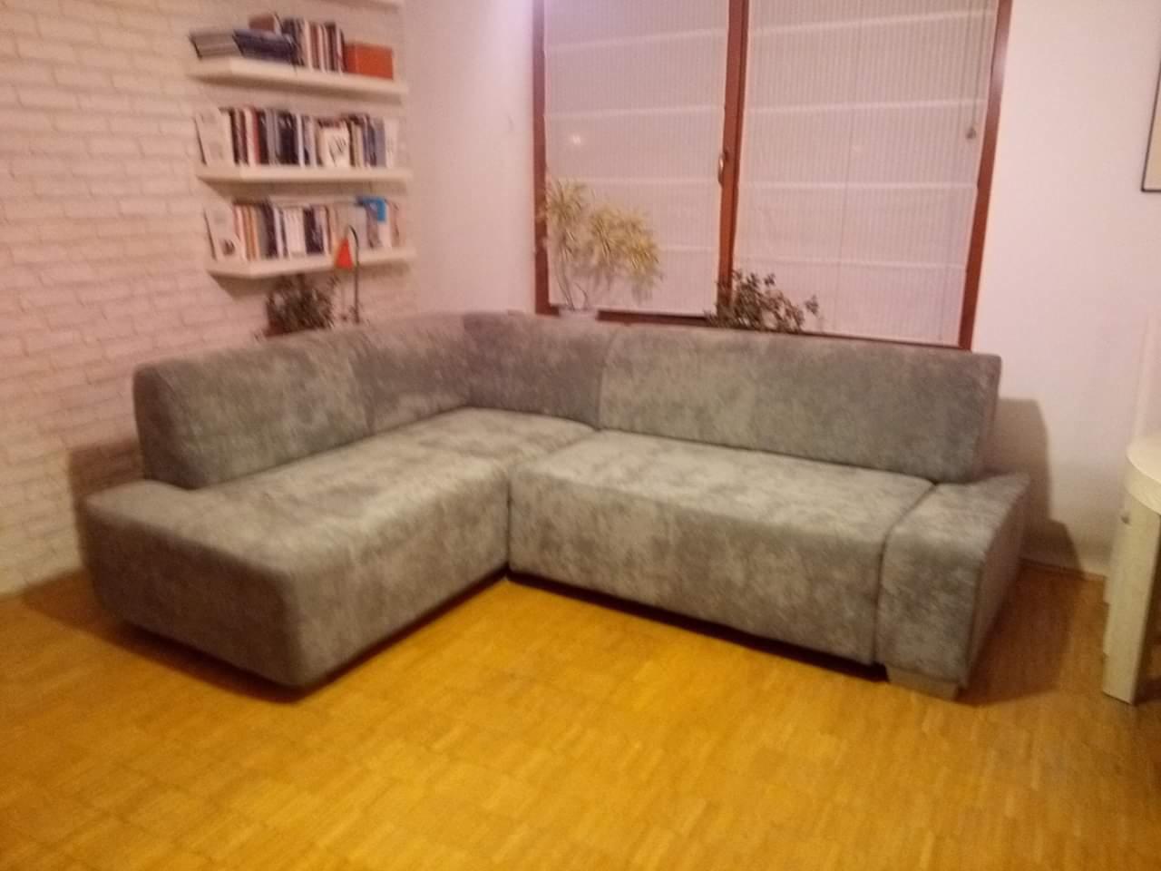 Szara narożna sofa z nową tapicerką w salonie z drewnianą podłogą, na tle ceglanej ściany z półkami na książki i okna.