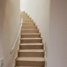 Schody wewnętrzne wyłożone płytkami imitującymi marmur, z metalową poręczą w folii ochronnej, przy białych ścianach.