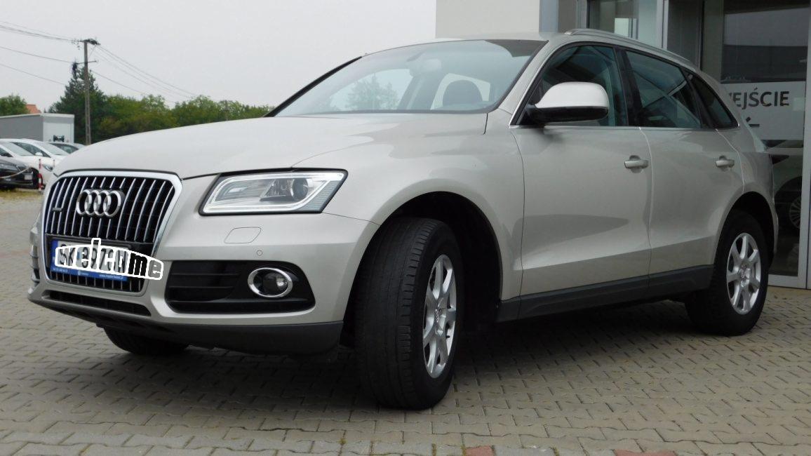 Srebrny SUV marki Audi, model Q5, zaparkowany przed budynkiem z napisem 'Wejście' na szklanych drzwiach, widoczne inne samochody w tle.