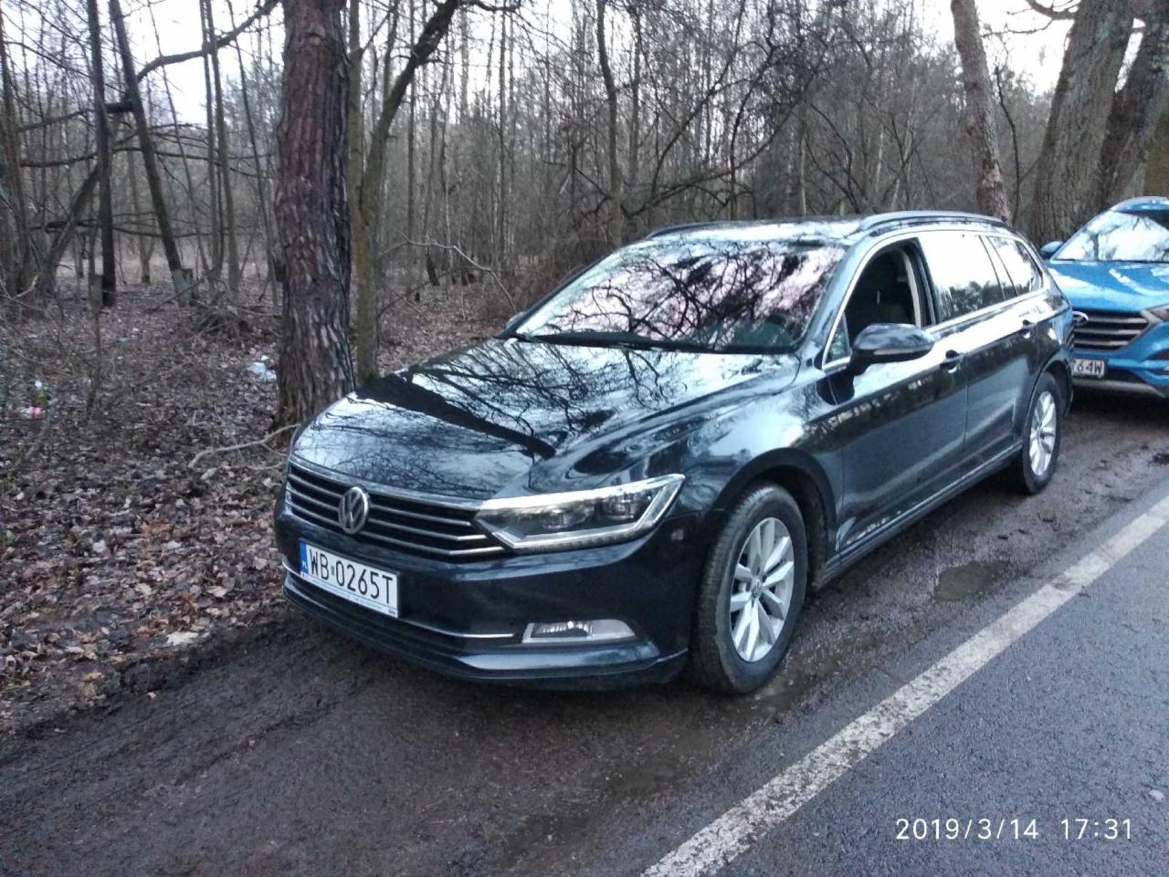 Czarny samochód kombi marki Volkswagen zaparkowany na poboczu drogi, obok lasu, z widoczną tablicą rejestracyjną WB 0265T i odbiciem drzew w szybie.