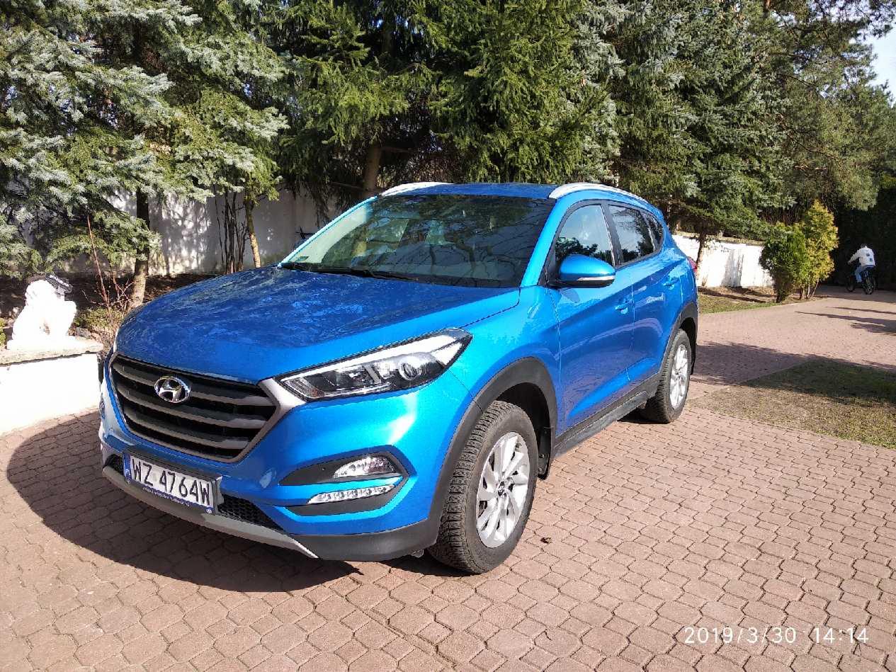 Błękitny Hyundai Tucson zaparkowany na brukowanej nawierzchni, z widocznymi drzewami w tle i osobą na rowerze w oddali.