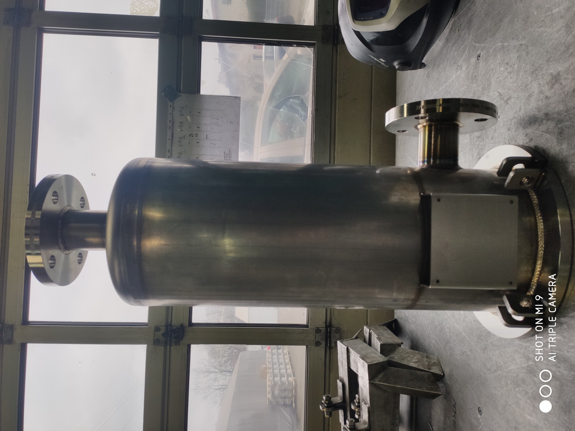 Metalowy zbiornik z przyspawanymi kołnierzami, leżący na stole warsztatowym w hali, widoczna maska spawalnicza w tle.