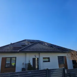 Biały dom jednorodzinny z ciemną dachówką i panelami fotowoltaicznymi na dachu, widok z poziomu ogrodzenia w słoneczny dzień.