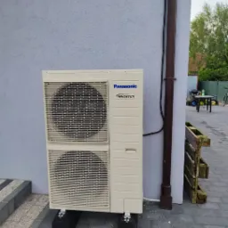 Zewnętrzna jednostka pompy ciepła Panasonic Inverter zamontowana na czarnych podstawach na szarej kostce brukowej, obok brązowa rynna spustowa i drewniana paleta.