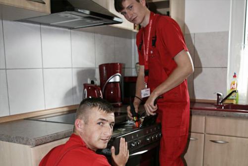 Dwóch mężczyzn w czerwonych kombinezonach instaluje płytę grzewczą w kuchni z białymi płytkami i beżowymi szafkami.