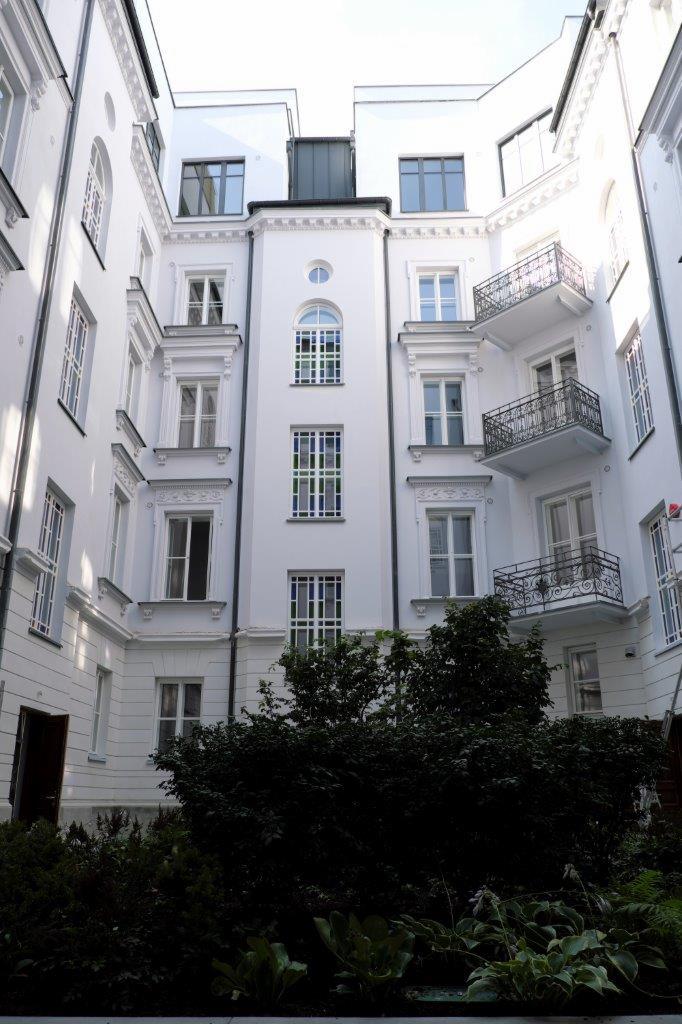 Wewnętrzny dziedziniec kamienicy z białymi elewacjami, ozdobnymi balkonami i oknami, częściowo zasłonięty zielenią.