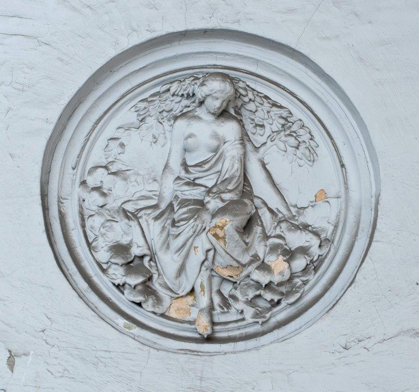 Okrągła, gipsowa płaskorzeźba przedstawiająca siedzącą, półnagą kobietę pod drzewem, umieszczona na popękanej, jasnej ścianie.