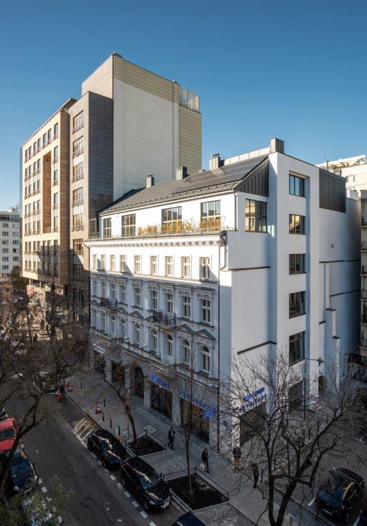 Elewacja odnowionej kamienicy z dobudowaną nowoczesną częścią, widok z góry na ulicę i zaparkowane samochody w słoneczny dzień.