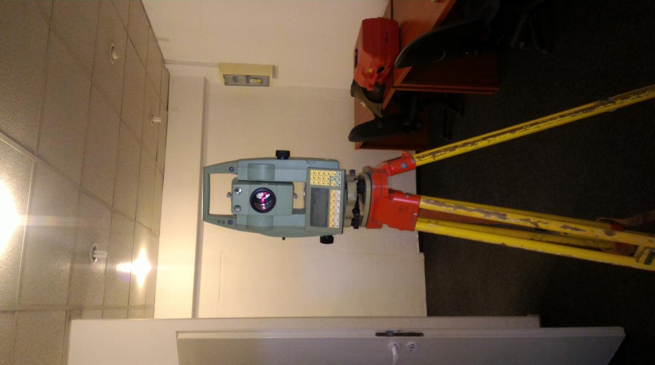 Teodolit na żółtym statywie z czerwonymi elementami, ustawiony w pomieszczeniu z kasetonowym sufitem i lampami.