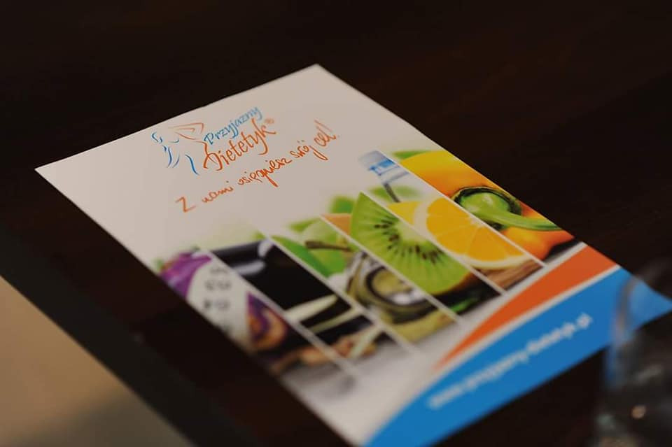 Ulotka reklamowa z logo i hasłem 'Przyjazny Dietetyk. Z nami osiągniesz swój cel!', zawierająca zdjęcia owoców i warzyw, leżąca na ciemnym blacie.